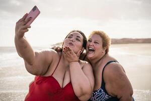 contento più dimensione donne assunzione autoscatto con mobile smartphone su il spiaggia - sovrappeso amici avendo divertimento durante vacanza tempo foto