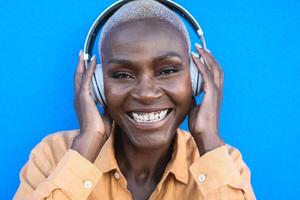 contento anziano africano donna avendo divertimento ascoltando musica con senza fili cuffie contro blu sfondo foto