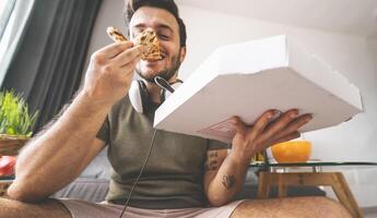 giovane uomo mangiare Pizza consegna a casa - contento tipo avendo pasto mentre giocando video Giochi nel vivente camera - cibo e gioventù persone divertimento concetto foto