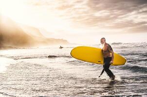 contento in forma anziano fare surf su tramonto tempo - sportivo maturo uomo avendo divertimento formazione con tavola da surf nel oceano - anziano salutare persone stile di vita e estremo sport concetto foto