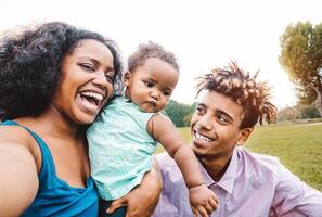 contento africano famiglia fare autoscatto foto con mobile Telefono nel un' pubblico parco all'aperto - madre e padre avendo divertimento con loro figlia durante un' fine settimana soleggiato giorno - amore e felicità concetto