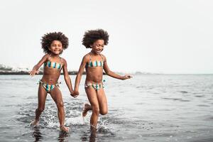 contento sorelle in esecuzione dentro acqua durante estate tempo - afro bambini avendo divertimento giocando su il spiaggia - famiglia amore e viaggio vacanza stile di vita concetto foto