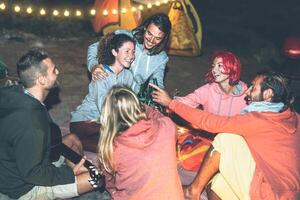 gruppo di amici applauso e tostatura con birra mentre campeggio con tenda Il prossimo il spiaggia - contento giovane persone avendo divertimento potabile nel campeggio con Vintage ▾ luci - viaggio e gioventù vacanze concetto foto