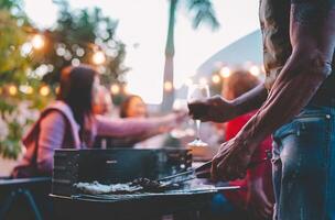 contento famiglia fare barbecue cena su tetto a notte - vicino su mano di tatuaggio uomo cucinando a bbq griglia all'aperto - concetto di cibo, persone stile di vita e estate fine settimana attività foto