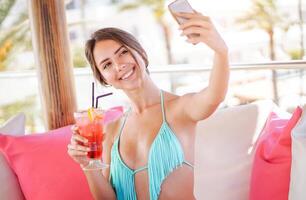 giovane bella donna assunzione un' autoscatto con mobile inteligente Telefono nel un' spiaggia club - attraente ragazza avendo divertimento potabile un' tropicale cocktail e spedizione fotografie su sua sociale reti durante sua vacanza