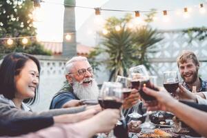 contento famiglia cenare e tostatura rosso vino bicchieri all'aperto - persone con diverso età e etnia avendo divertimento nel barbecue cena festa - paternità gioventù e anziano fine settimana attività concetto foto