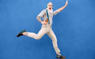 anziano pazzo uomo salto mentre ascoltando musica con senza fili cuffie - fricchettone vecchio tipo avendo divertimento danza all'aperto - persone felicità e anziano tecnologia dipendente concetto - blu sfondo foto