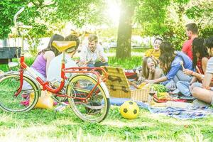 gruppo di un' lotto di persone avendo un' gustoso picnic mangiare e potabile rosso vino nel un' parco all'aperto - contento amici festeggiare un' soleggiato giorno condivisione bevande e Alimenti - tradizionale pasto fuse con natura foto