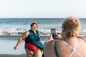 contento più dimensione donne avendo divertimento fabbricazione fotografie su il spiaggia con mobile smartphone - sovrappeso fiducioso persone stile di vita e tecnologia concetto