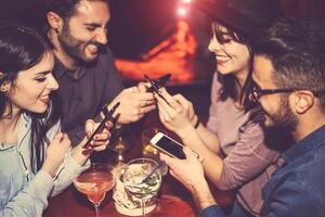 contento amici utilizzando mobile Telefono e avendo divertimento con cocktail nel un' jazz bar - giovane persone dipendente per nuovo smartphone tecnologia - concetto di gioventù, cellulare e stile di vita - messa a fuoco su giusto maschio mano foto