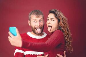 contento pazzo coppia assunzione autoscatto mentre fare sciocco viso con mobile smartphone telecamera - giovane persone festeggiare Natale vacanze - amore relazione, natale e tecnologia tendenze concetto foto