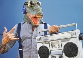 pazzo anziano uomo avendo divertimento indossare t-rex maschera mentre ascoltando per musica con cuffie e Vintage ▾ Boombox stereo all'aperto foto