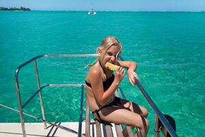 un' poco ragazza nel un' nero costume da bagno mangia ananas su vacanza su un' yacht a mare foto