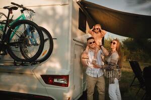 un' giovane famiglia con bicchieri è in piedi Il prossimo per loro mobile casa. papà, mamma e figlia insieme vicino il camper foto