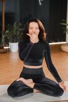 un' donna nel un' nero tuta da ginnastica si siede su un' yoga stuoia nel un' loto posizione nel il Palestra foto