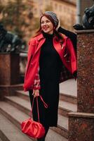 un' bellissimo elegante donna vestito nel un elegante rosso cappotto con un' elegante rosso borsetta nel il autunno città foto