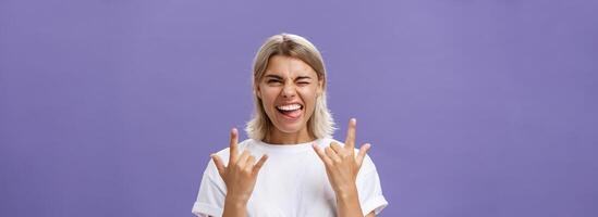 andando roccia. ritratto di gioioso contento bella vista elegante donna con biondo medio taglio di capelli strizzando l'occhio sorridente e attaccare su lingua mentre mostrando rock n roll gesto con tutti e due mani al di sopra di viola parete foto