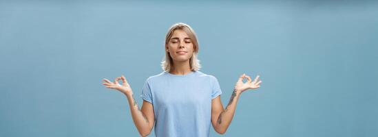 ascoltando per interno se stesso durante meditazione. calma contento e rilassato attraente abbronzato bionda femmina con tatuaggi su braccia in piedi nel loto posa al di sopra di blu parete con zen gesto chiuso occhi fare yoga foto