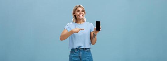 Guarda a Questo divertente foto. intrattenuto attraente contento donna con giusto capelli nel casuale maglietta e denim pantaloncini mostrando smartphone a telecamera puntamento a dispositivo schermo sorridente a grandi linee al di sopra di blu parete foto