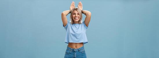 tempo per relax. ritratto di contento giocoso carino bionda ragazza nel di moda maglietta con trafitto pancia e tatuaggio sorridente con gioia Tenere palme su testa piace coniglietto orecchie avendo divertimento al di sopra di blu sfondo foto