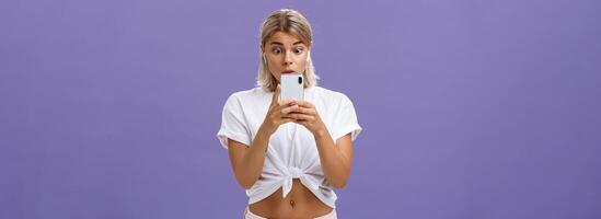 ragazza Guardando emozionante video su sociale media nel senza fili auricolari Tenere smartphone vicino viso popping occhi a smartphone schermo guardando a aggeggio con interesse e curiosità al di sopra di viola sfondo foto