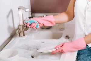 casalinga ragazza nel rosa guanti lavaggi piatti di mano nel il Lavello con detergente. il ragazza pulisce il Casa e lavaggi piatti nel guanti a casa. foto