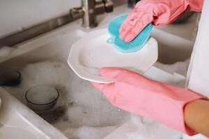 casalinga ragazza nel rosa guanti lavaggi piatti di mano nel il Lavello con detergente. il ragazza pulisce il Casa e lavaggi piatti nel guanti a casa. foto