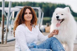 un' contento donna con un' grande bianca cane si siede su un' molo di il mare a tramonto foto