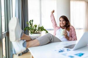 giovane bella donna d'affari nel il ufficio seduta nel ufficio sedia, mani su e con sua piedi su su un' scrivania godendo caffè foto