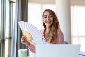 stanco surriscaldato giovane donna hold onda fan soffrire a partire dal calore sudorazione interno opera su il computer portatile a casa ufficio, infastidito donna sentire scomodo caldo estate tempo metereologico problema no aria condizionatore concetto foto