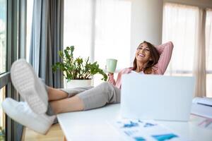 giovane bella donna d'affari nel il ufficio seduta nel ufficio sedia, mani su e con sua piedi su su un' scrivania godendo caffè foto