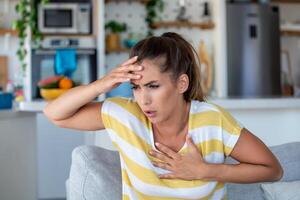 stressato giovane donna sensazione dolore e toccante il petto soffrire a partire dal angoscia malattia a casa mentre avendo cuore attacco, infarto foto