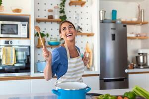 eccitato donna cantando e danza nel moderno cucina a casa, contento donna Tenere spatola come microfono, ballare, ascoltando per musica, avendo divertimento con stoviglie, preparazione prima colazione foto