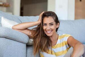 contento giovane donna seduta su divano a casa e guardare a telecamera. ritratto di bellissimo donna sorridente e rilassante durante estate. foto