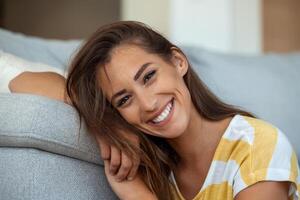 contento giovane donna seduta su divano a casa e guardare a telecamera. ritratto di bellissimo donna sorridente e rilassante durante estate. foto