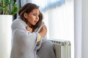 indisposto donna locatario nel coperta sedersi nel freddo vivente camera mano su vecchio radiatore.soffrire a partire dal mancanza di calore . malsano giovane donna lotta a partire dal freddo congelare a casa. no riscaldamento concetto. foto