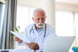 grave vecchio maturo professionale maschio medico utilizzando il computer portatile computer nel ospedale ufficio avendo medico seminario web addestramento, scrittura nel assistenza sanitaria rapporto, consulenza paziente in linea a telemedicina incontro. foto