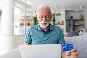 maturo uomo utilizzando computer portatile, Tenere plastica credito o addebito carta, anziano grigio dai capelli cliente fabbricazione sicuro Internet pagamento, shopping o navigazione in linea bancario servizio, entrata informazione foto