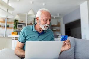 maturo uomo utilizzando computer portatile, Tenere plastica credito o addebito carta, anziano grigio dai capelli cliente fabbricazione sicuro Internet pagamento, shopping o navigazione in linea bancario servizio, entrata informazione foto