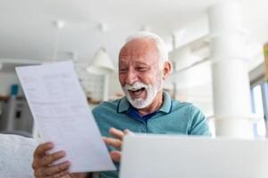 sorpreso ridendo contento vecchio maturo pensionato uomo guardare attraverso carta documento, sensazione eccitato analizzando finanziario informazione, ottenere le tasse rimborso o banca prestito approvazione a casa. foto