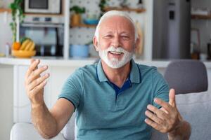 sorridente anziano uomo onda per telecamera avendo video chiamata su computer portatile, contento anziano maschio sedersi su divano a casa parlare utilizzando moderno tecnologie e senza fili connessione foto