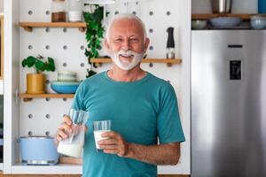 anziano uomo potabile un' bicchiere di latte con un' contento viso in piedi e sorridente. bello anziano uomo potabile un' bicchiere di fresco latte nel il cucina foto