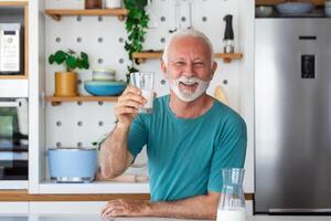 anziano uomo potabile un' bicchiere di latte con un' contento viso in piedi e sorridente. bello anziano uomo potabile un' bicchiere di fresco latte nel il cucina foto