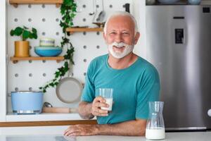 anziano uomo potabile un' bicchiere di latte con un' contento viso in piedi e sorridente. bello anziano uomo potabile un' bicchiere di fresco latte nel il cucina foto