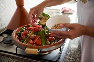 avvicinamento della casalinga mani impilamento fresco verdure nel un' stoviglie a partire dal argilla, preparazione cucinando tagine nel il cucina foto