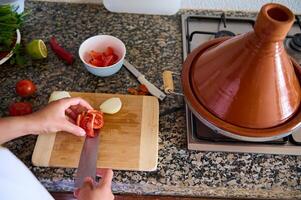 avvicinamento Visualizza a partire dal sopra di un' casalinga chopping pomodori su un' taglio tavola. un' argilla tajine pentola su un' acciaio stufa foto