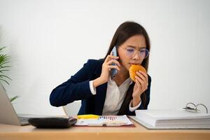 occupato e stanco donna d'affari mangiare pane e latte per pranzo a il scrivania ufficio e Lavorando per consegnare finanziario dichiarazioni per un' capo. oberati di lavoro e malsano per pronto pasti, bruciato concetto. foto