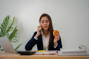 occupato e stanco donna d'affari mangiare pane e latte per pranzo a il scrivania ufficio e Lavorando per consegnare finanziario dichiarazioni per un' capo. oberati di lavoro e malsano per pronto pasti, bruciato concetto. foto