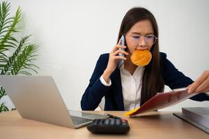 occupato e stanco donna d'affari mangiare pane e latte per pranzo a il scrivania ufficio e Lavorando per consegnare finanziario dichiarazioni per un' capo. oberati di lavoro e malsano per pronto pasti, bruciato concetto. foto