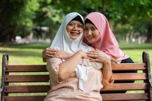 musulmano attento badante o infermiera assunzione cura di il paziente nel il ospedale parco. contento musulmano madre nel hijab abbracciare figlia. concetto di risparmi e anziano Salute assicurazione, un' contento famiglia foto